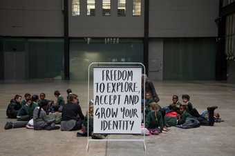 A group of young people in school uniform sit in a circle on the floor at Tate Modern. A sign stands in front of them, reading “Freedom to explore, accept and grow your identity.”
