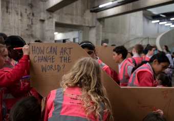A group of young people in pink high-vis vests write onto cardboard structures. On the cardboard is the question, ‘How can art change the world’?