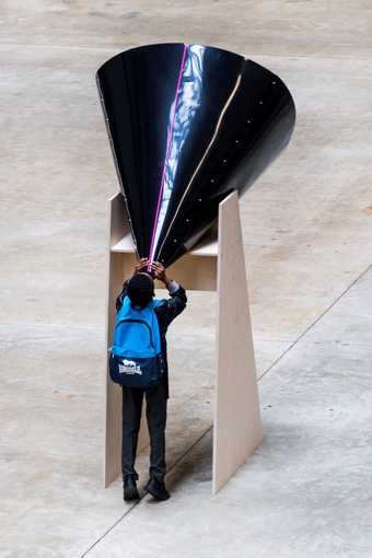 A young person looks up through a large, black cone in the gallery.