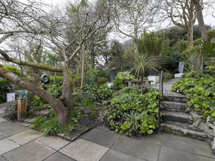 The garden on a cloudy day. It is filled with lush green plants, tall trees and metal sculptures. The ground is paved with stone and there are uneven stone steps with a handrail on the right.