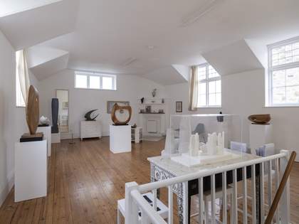 The room is bright, with windows, white walls and a wooden floor. There are plinths where large sculptures sit. Some items hang from the walls, like paintings and a mirror.
