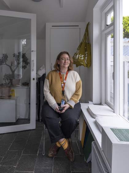 A smiling Visitor Assistant sits on a stool by a large bright window.