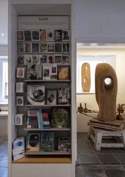 A tall shelving cabinet displaying a large range of colourful books and postcards. A sign at the top reads ‘SHOP’. To the right of the cabinet there is a wooden stool where a large wooden sculpture sits.