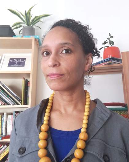 A mid view of a woman wearing a large beaded necklace, jacket and blue top.  There are bookshelves behind her.