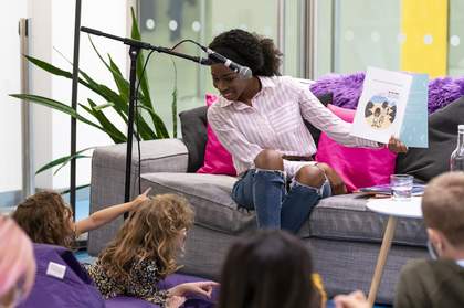 A person with a microphone on a stand sitting on a sofa shows children a picture from a storybook