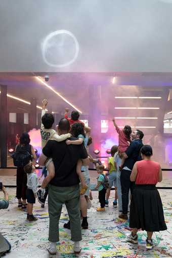 A family at a disco at Tate Modern.  The figures are facing away and there is a large smoke ring in the air, as well as neon lights.