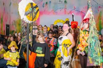 A large group of children and adults wearing colourful handmade outfits and bright face paint, with a paint splattered backdrop