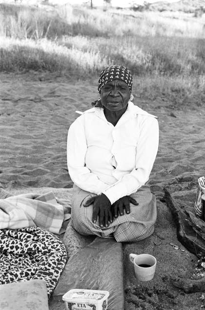 Emily Kam Kngwarray kneeling on blankets, with sand and grass behind her, and a cup of tea in front of her.