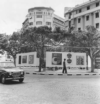 A black-and-white photograph of a cobbled street corner with artworks hanging on boards in the middle of the square