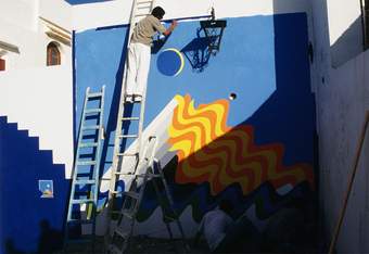 A photograph of the artist Mohamed Melehi at the top of a step ladder working on an abstract blue, yellow and orange mural