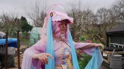 A person dressed in a cosplay costume. They are wearing a pink and blue costume, a light pink dragon head with yellow and black eyes as a hat, with pink gauze running down their back and blue gauze draped from the eyes like tears.