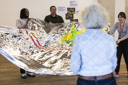 A group of teachers use a large piece of fabric made out of foil blankets to toss crumpled pieces of tissue paper up into the air.