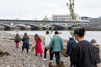 8 people walking away along the Thames riverbank, some holding branches, with Blackfriars bridge in the distance.