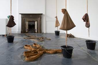 An artwork installation including two clay sculptures and four buckets on the floor with four bags of clay suspended above.