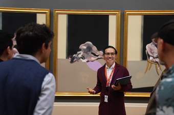Person smiling and holding a laptop in one arm and wearing a maroon top and tate lanyard, stood in front of three large paintings in gold frames. They are smiling and looking to people whose backs are blurred in the foreground.