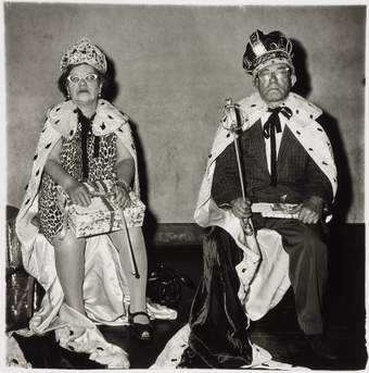 Two people sitting down on chairs dressed as royalty with crowns, robes and holding sceptres