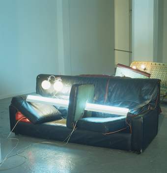 Sculpture of a dishevelled leather sofa with a long, thin fluorescent light piercing through the middle of one of the cushions on it's side in the centre with a wall light on one-side