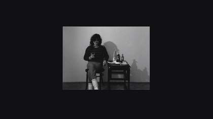 A black and white film still of a person sitting on a chair smiling, with a table next to them with bottles and a plate on it