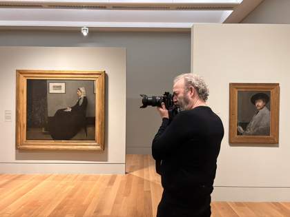 A side view of a man wearing black in a gallery with a camera held up to his face