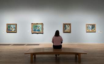 A woman sat on a bench facing a white wall with a line of paintings.