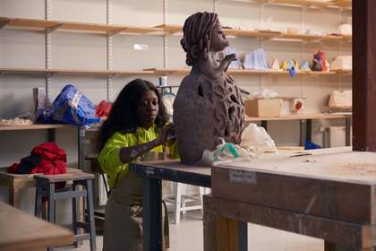 Image shows artist Josie Ko standing at a work bench in a studio space, with shelving across the walls behind her. She is working on a clay sculpture of a female figure.