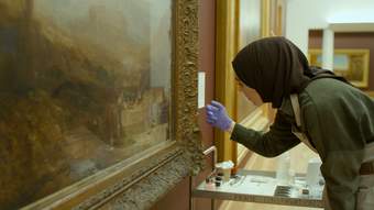 A person wearing blue gloves in an art gallery tapping on a picture frame of a painting