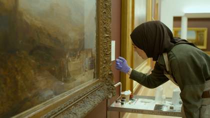 A person wearing blue gloves in an art gallery tapping on a picture frame of a painting