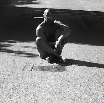 A man with his legs crossed sits on a gravel floor, the photograph is shot in black and white.