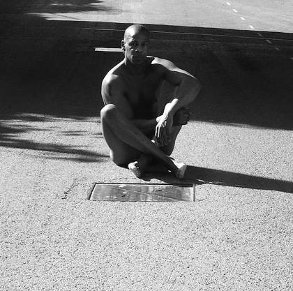 A man with his legs crossed sits on a gravel floor, the photograph is shot in black and white.