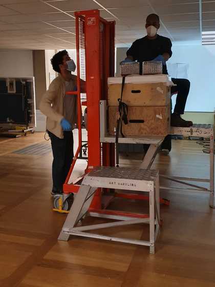 An individual operating a raised platform and another person sitting on the platform with their gloved hands placed on two glass lamps