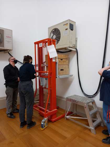 Individuals standing in front of a raised platform that sits underneath a wall-mounted fan