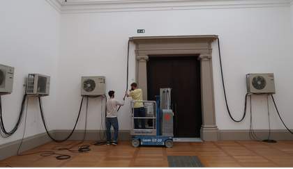 Two individuals holding up a long wire that connects several wall-mounted fans together and runs along the top of a large doorway flanked by pillars