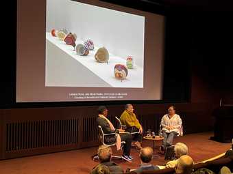 Three people sit on a stage and talk, with a large image of colourful ceramic pots projected on a screen behind them, and an audience watching