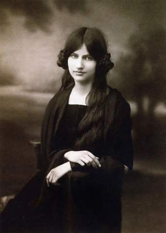 Black-and-white studio portrait of Jeanne Hébuterne seated in front of a rural landscape backdrop, looking directly into the camera. She wears dark clothing against which her skin appears bright and luminous.