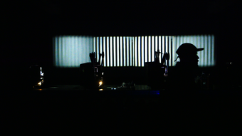 A very dark room; white vertical lines are projected onto a wall. Somebody's head, in the way of the projector, appears silhouetted against the white lines. Other objects are just visible: perhaps instruments, music stands or microphones