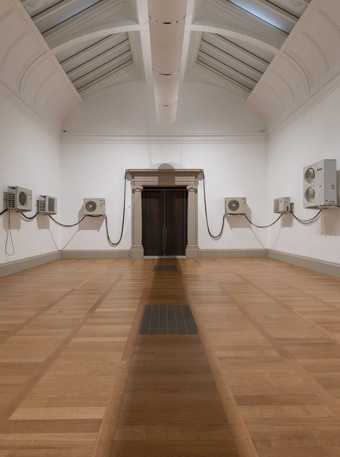 Air-conditioning units mounted on the wall of an art gallery, evenly spaced and connected to each other by wires