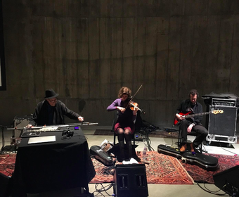 Three people sitting in a row in front of the concrete wall of the Tanks, each playing a stringed instrument: the long string drone, violin and guitar