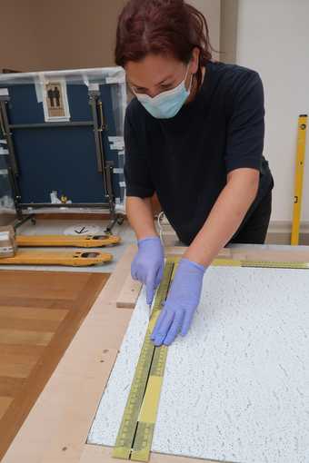 Gloved hands holding a knife against a ruler on top of a large flat tile and cutting into it