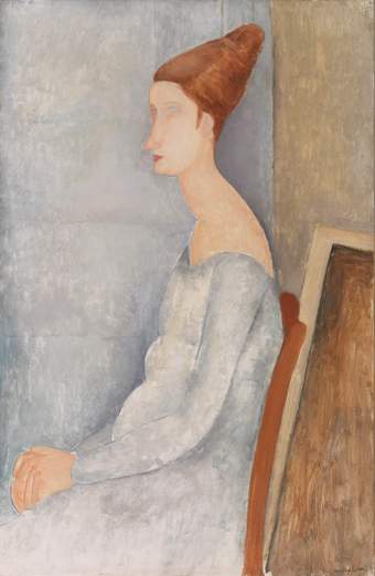 Hébuterne seated in profile, hands in her lap, head turned toward the viewer. Shoulders are rounded, emphasised by a pale blue-grey dress cut low. Her red hair is styled up in a cone shape. The dress blends into a similarly coloured wall behind.
