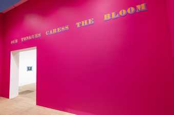 A bright pink wall with the words ‘Our tongues caress the bloom’ written on it in large letters, above an archway that leads into a white gallery space