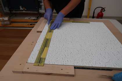 Gloved hands holding a knife against a ruler on top of a large flat tile and cutting into the tile