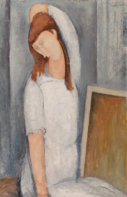 Portrait of Hébuterne, sitting with legs folded beneath her, one arm reaching upwards around her head. She looks down, red hair loose. Her dress is pale blue-grey against the darker blue-grey background. A shape next to her, like the back of a canvas