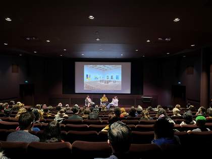 An audience sits in an auditorium watching three people talk on a stage
