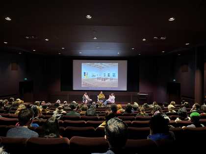 An audience sits in an auditorium watching three people talk on a stage