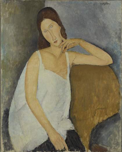 Three-quarter portrait of Hébuterne. She sits in a dark yellow armchair, one arm propped on the top of the chair, a finger of one hand against her cheek, her head tilted to one side, in a playful or flirtatious gesture. She wears a white sleeveless dress.