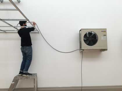 An individual standing on a small platform and holding up a long wire that connects to a fan mounted on the wall
