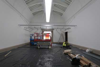 A view of an art gallery with a metal grid installed horizontally across it above head height, and black plastic sheeting, cardboard boxes and industrial trolleys underneath it