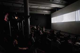 A group of people sit in a darkened room looking at an image of vertical strips of white light that is projected onto the wall