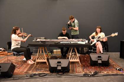 Three musicians play a violin, a long guitar-like instrument and a bass guitar, while being filmed with a camera