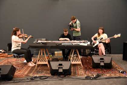 Three musicians play a violin, a long guitar-like instrument and a bass guitar, while being filmed with a camera